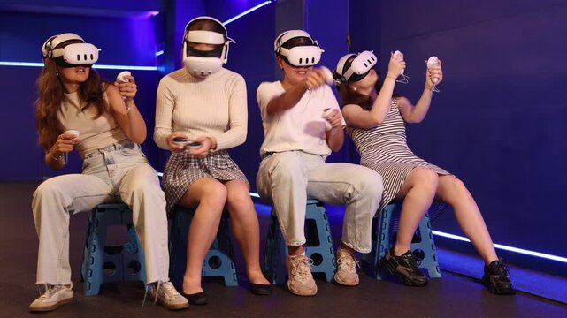 Group of excited young girls sitting with handheld controllers in hands engaged in virtual world experiencing through VR glasses covering face. Concept of immersion and interactivity in game