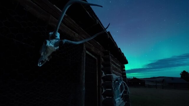 Slider move past an old log cabin with an elk skull and antlers as green northern lights glow over a ranch, Montana, time lapse