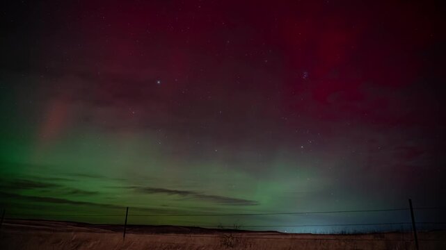 Intense red and green northern lights swirling through clouds above a fence line on the open plains, Montana, time lapse
