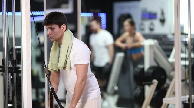 During workout, guy with towel on neck performs grueling exercises and pumps muscles of arms and upper torso on crossover exercise machine, pull rope and make work out.