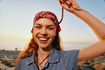 smiling woman with bandana and braided hair wearing denim shirt enjoying sunny beach outdoors with...