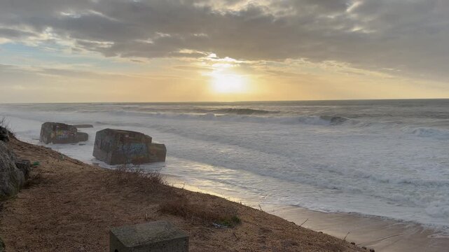 4K static shot of historical World War 2 concrete bunkers on a sandy beach at sunset. Intense Atlantic Ocean waves crash against the graffiti-covered ruins under a dramatic, moody, overcast cloudy sky