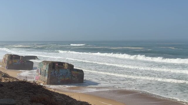4K static shot of historic World War II bunkers on a sandy beach, covered in colorful graffiti. Waves from the Atlantic Ocean crash against the concrete ruins. Cinematic coastal landscape in Labenne, 