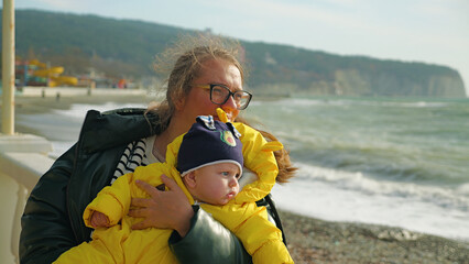 A mother wearing glasses tenderly holds her infant son, who is bundled in a vibrant yellow puffer...