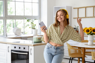 Cool young woman with mobile phone listening to music and dancing in kitchen