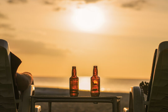 Two bottles of beer are on the table at sunset on the beach - Two friends or lovers are watching the sunset sitting in chairs back view. The concept travel and relaxing holidays with relaxing drinks
