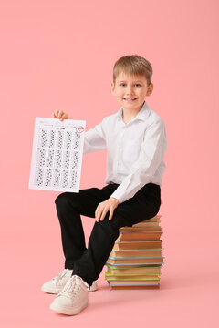 Happy schoolboy with good results of school test sitting on books against pink background