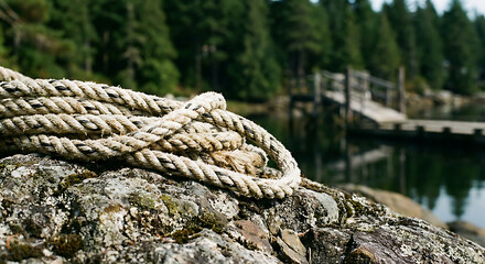 Fototapeta premium thick braided rope coiled on a mossy rock by a serene lake with a rustic wooden dock and green forest.
