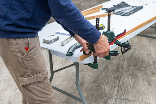 Man in casual clothes clamps a wooden plank to a folding worktable, focused on precise woodworking and home repair during a diy renovation project indoors