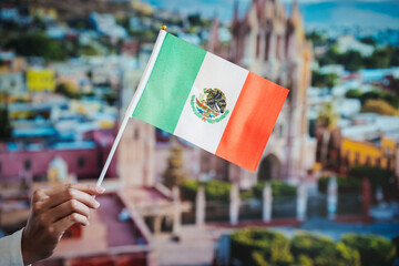 Hand holding mexican flag on small pole, blurred historic cityscape and cathedral in background,...