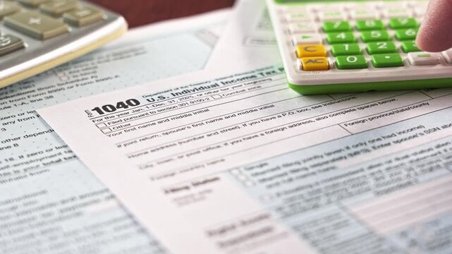 Close-up view of a person's finger pressing keys on a calculator. Financial documents, specifically the american 1040 tax form, in the background
