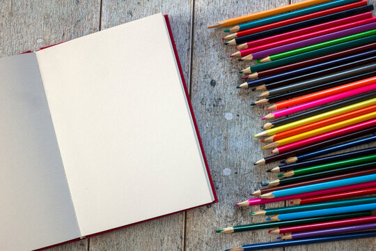 Top view of a blank sketchbook surrounded by colorful pencils on rustic wooden background. Perfect for DIY, creative hobby, art projects, drawing, sketching, and school supplies concept.