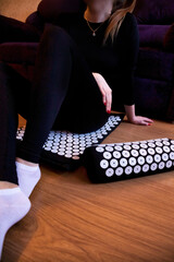 A woman is sitting on the floor in her living room, using an acupressure mat and pillow for self massage. She is relaxing and focusing on her well-being at home.