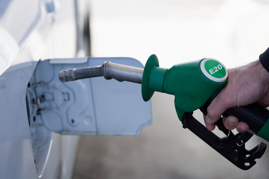 man refilling car with e20 biofuel at gas station, hand holding green fuel nozzle with e20 label close up, white vehicle parked outdoors, concept of oil industry, energy sector, fuel economy