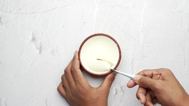 Taking spoonful of thick creamy yogurt from traditional clay pot on white textured surface in slow motion