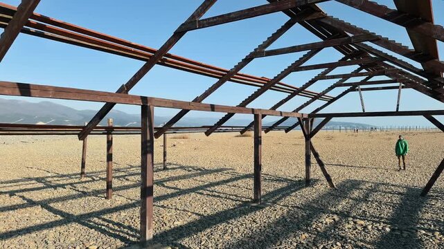 Authentic Front View Of Bearded Male Traveler In Green Hoodie With Backpack Approaching And Passing By Camera Through Abandoned Wooden Skeletal Frame On Desolate Pebble Beach Against Blue Sky