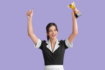 Happy young chambermaid with gold cup on lilac background
