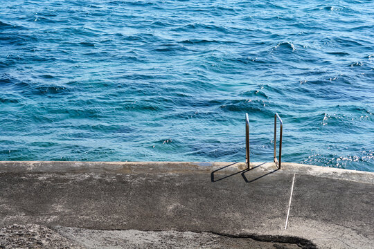 A set of stainless steel handrails for a sea access ladder stands on a stone promenade by the rippling blue waters of the Adriatic Sea.