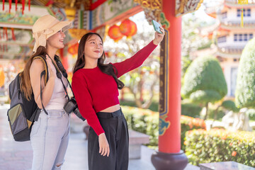 Obraz premium Two happy asian women tourists taking selfie with smartphone at Chinese temple, Young backpacker friends waving to video call on summer vacation holiday, Culture travel and technology concept