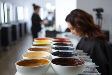 colorist preparing multiple bowls of different hair dye shades, client in background, minimalist beauty studio, professional lighting setup