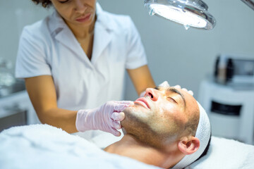 Fototapeta premium esthetician performing professional acne treatment on male client, focus on inflamed pimple area, client lying on treatment bed, clean spa interior, bright clinical lighting