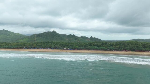 Aerial view of a stunning tropical sandy beach on java island. Turquoise ocean waves washing ashore, backed by lush green mountains and an overcast sky