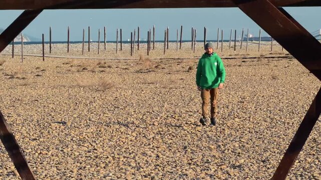 Authentic Man Walking Toward Camera Through Wooden Arch On Pebble Beach With Distant Cargo Ships Golden Hour Slow Motion