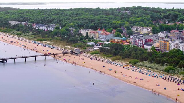 An aerial view shows the Binz pier on R&uuml;gen stretching into the Baltic Sea, lined with a busy sandy beach and classic resort architecture.