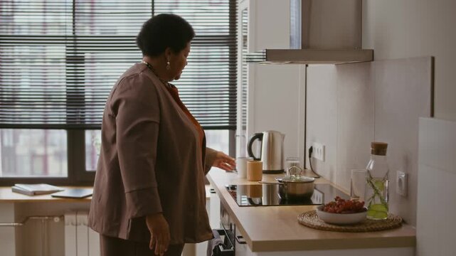 Medium slowmo shot of mature Black woman using induction stove while cooking porridge in small saucepan, preparing breakfast and drinking tea or coffee from mug in modern kitchen