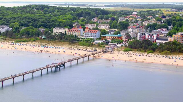 An aerial view shows the Binz pier on R&uuml;gen stretching into the Baltic Sea, lined with a busy sandy beach and classic resort architecture.