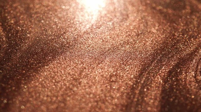 Sparkling view of rose gold paint revealing a specialized sandy texture with deep swirling ridges while light moves across causing glitter particles to shine intensely on dark background