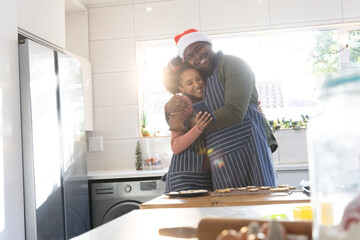 African American dad and daughter in striped aprons hugging at counter holding phone by cookie tray
