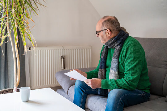 Man in scarf checking heating bill on sofa indoors during cold weather
