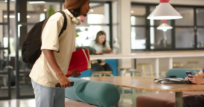 African American man entering left in study lounge, placing red folder, talking to peer, copy space