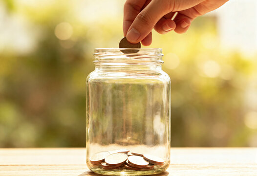 A hand is gently placing a coin into a glass jar containing more coins, set against a blurred, sunlit background, symbolizing saving money and financial planning concepts