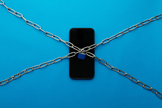 A black smartphone secured with metal chains and a blue padlock on a bright blue background. Concept of digital addiction, phone dependency, social media restraint, and losing freedom to technology.