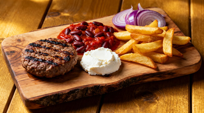 Authentic Serbian pljeskavica with kajmak cheese, prebranac beans and fries served on rustic wooden board.