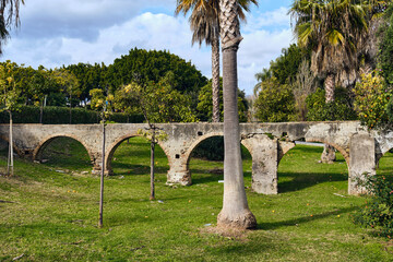 An old arched bridge ruins stand in a green park with palm trees under a cloudy sky Outdoor architecture and landscape photography.