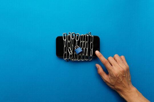 A hand reaching to touch a smartphone wrapped in chains and padlocked. Conceptual image for phone dependency, digital addiction craving, loss of control, and the struggle of a digital detox journey.