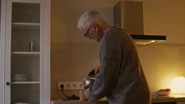 Medium shot of elderly Caucasian man with gray hair pouring water from elective kettle into mug in cozy modern kitchen and scrolling newsfeed on smartphone while enjoying tea in evening