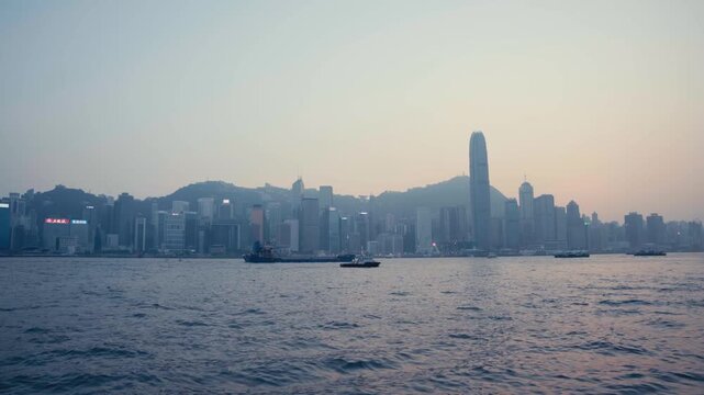 Wide static shot of Hong Kong skyline across Victoria Harbour with cargo ships on calm water and mist-shrouded skyscrapers including ICC tower during twilight dusk light