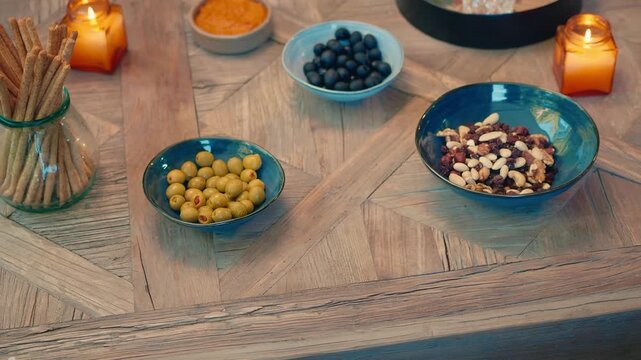 Bowls containing olives, nuts, and blueberries rest on a patterned wooden table accompanied by breadsticks and glowing amber candles ready for guests.
