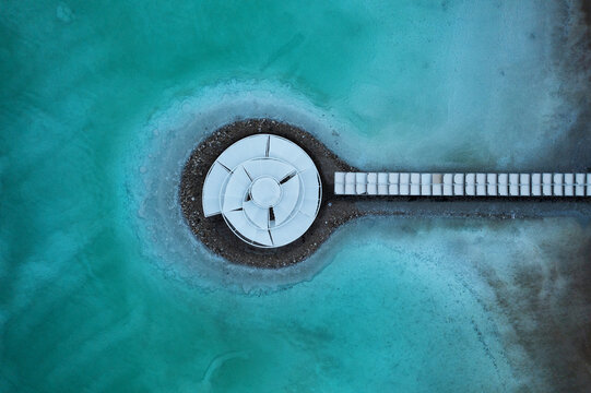 Aerial view of a circular structure with a walkway extending into turquoise waters, a serene contrast of geometric forms and natural hues, Arad, South District, Israel.