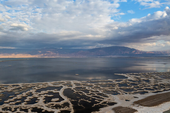 Aerial view of the Dead Sea's waters reflecting the cloudy sky and surrounding desert landscape, the salt formations contrasting with the dark water, Arad, South District, Israel.