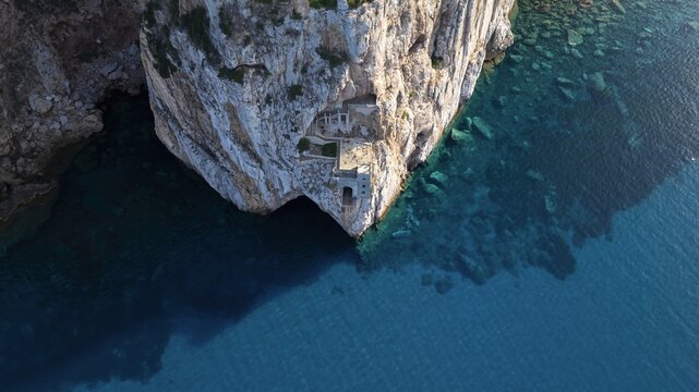 Aerial view of a rugged cliff face descending into the turquoise sea, revealing hidden architectural wonders carved into the stone, Masua, Sardegna, Italy.