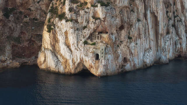 Aerial view of a rugged cliff face plunging into the deep blue sea, revealing an old building nestled within, Masua, Sardegna, Italy.