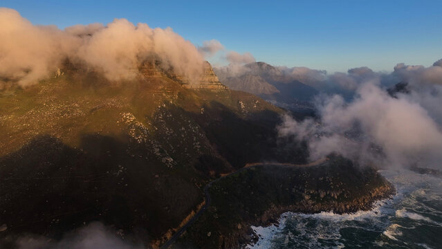 Aerial view of dramatic clouds clinging to the mountains, contrasting with the rugged coastline and crashing waves, Cape Town, Western Cape, South Africa.
