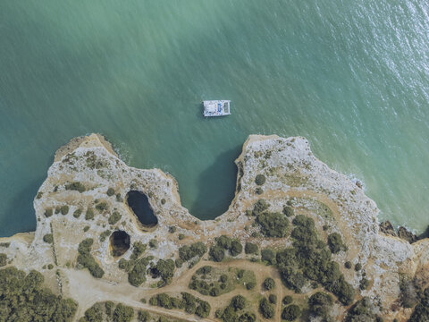 Aerial view of a boat floating on turquoise waters near rugged cliffs with sinkholes and sparse vegetation, Porches, Faro, Portugal.