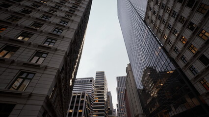 Naklejka premium low angle glass skyscraper canyon dusk, brick facades and mirrored towers framing narrow street, warm office lights glowing through windows, architect sketching