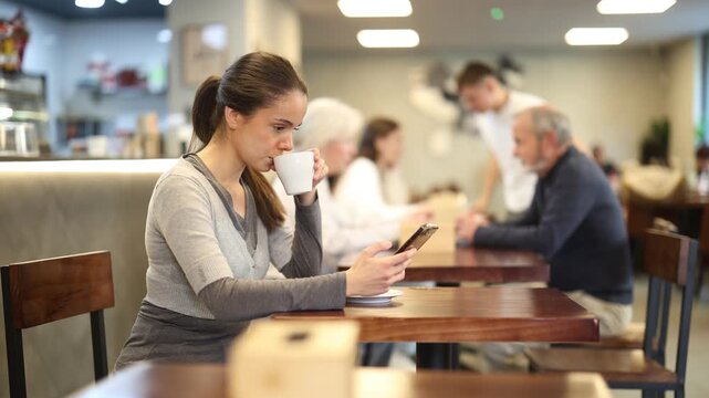 Girl in cafe drinks coffee and using mobile phone, scrolls through social media feed, chats online, calls friend, talks on phone. Free Wi-Fi zone, Internet cafe.